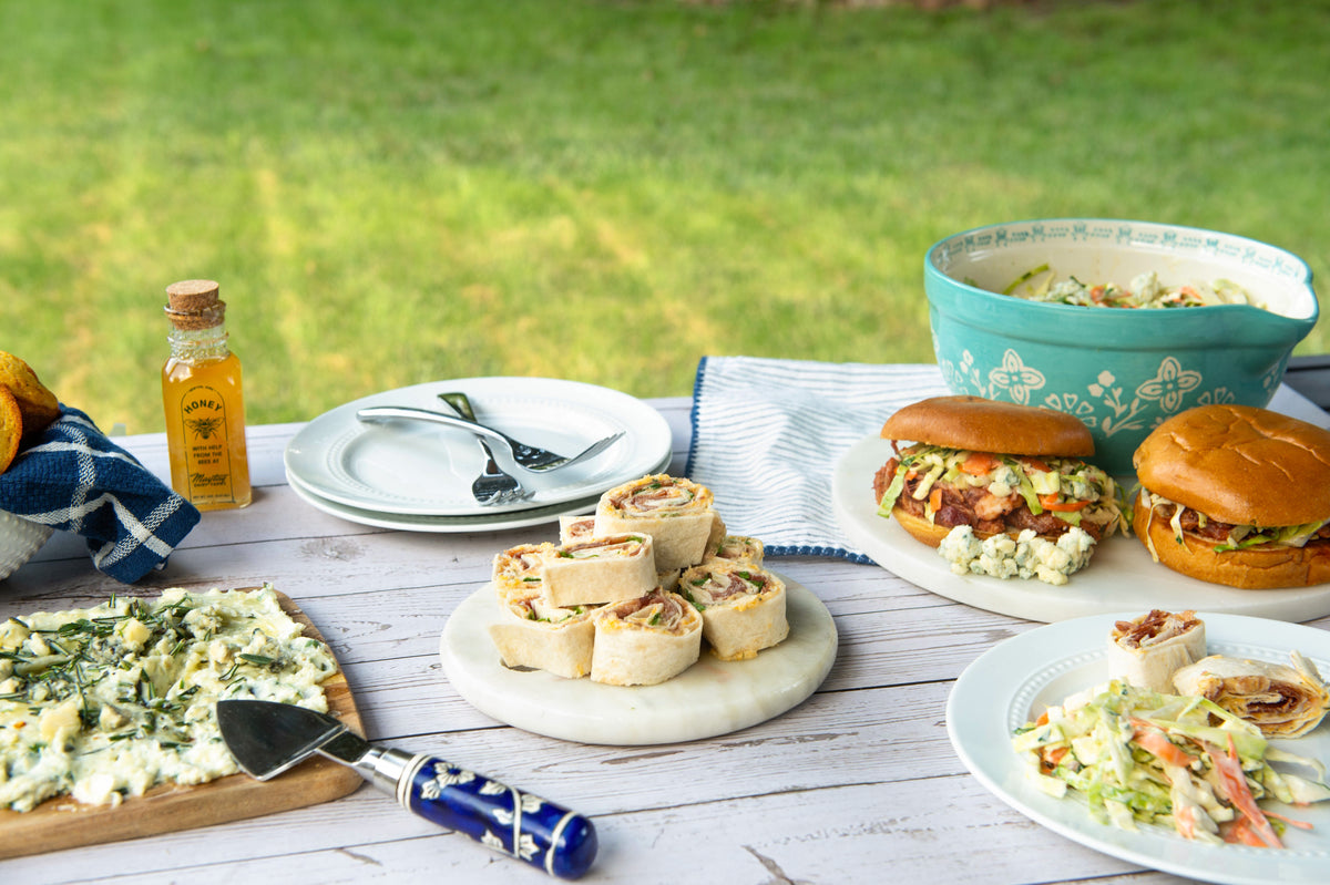 Outdoor picnic-style food spread on a white wooden table with a blurred green lawn background. Visible items include two sliders on buns, a platter of stuffed rolls, a side salad, a cheese spread on a board with a serving utensil, stacked plates, a small bottle of honey, and a turquoise bowl.